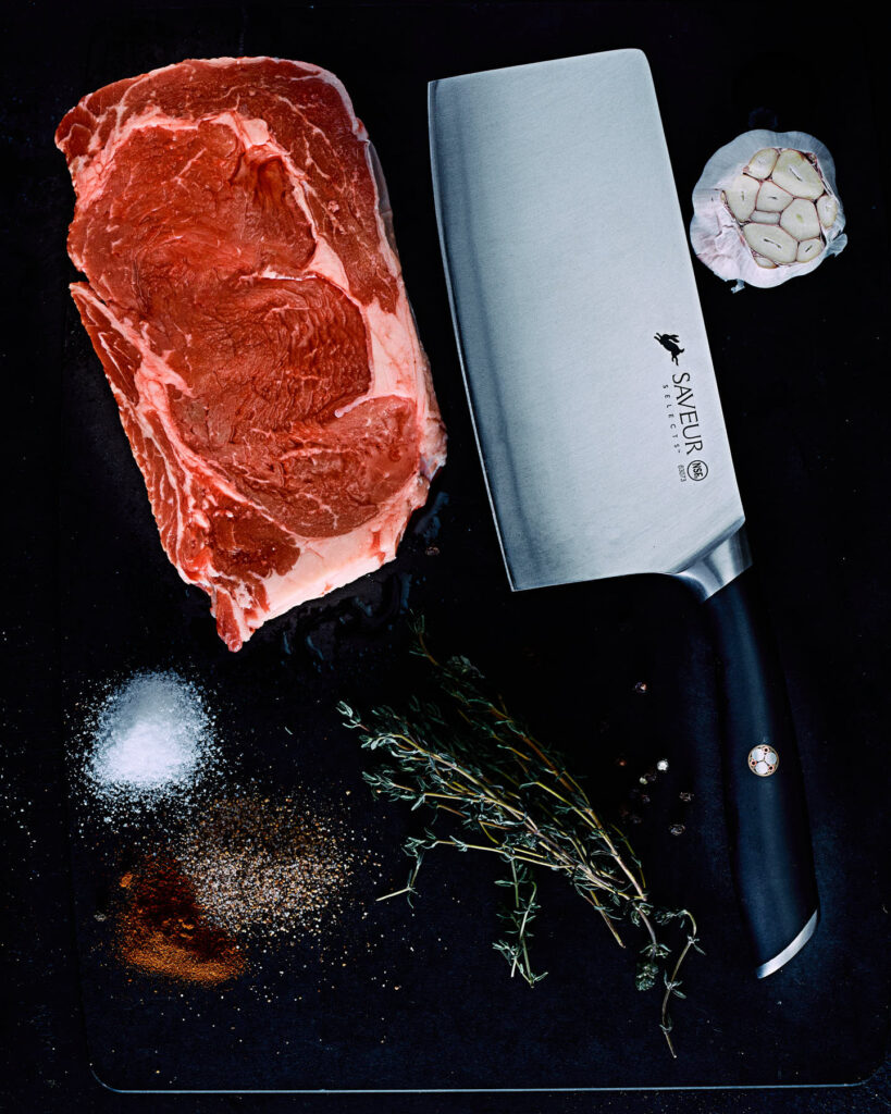 Filet mignon steak with a meat cleaver, salt, pepper, garlic and rosemary on top of a black cutting board.