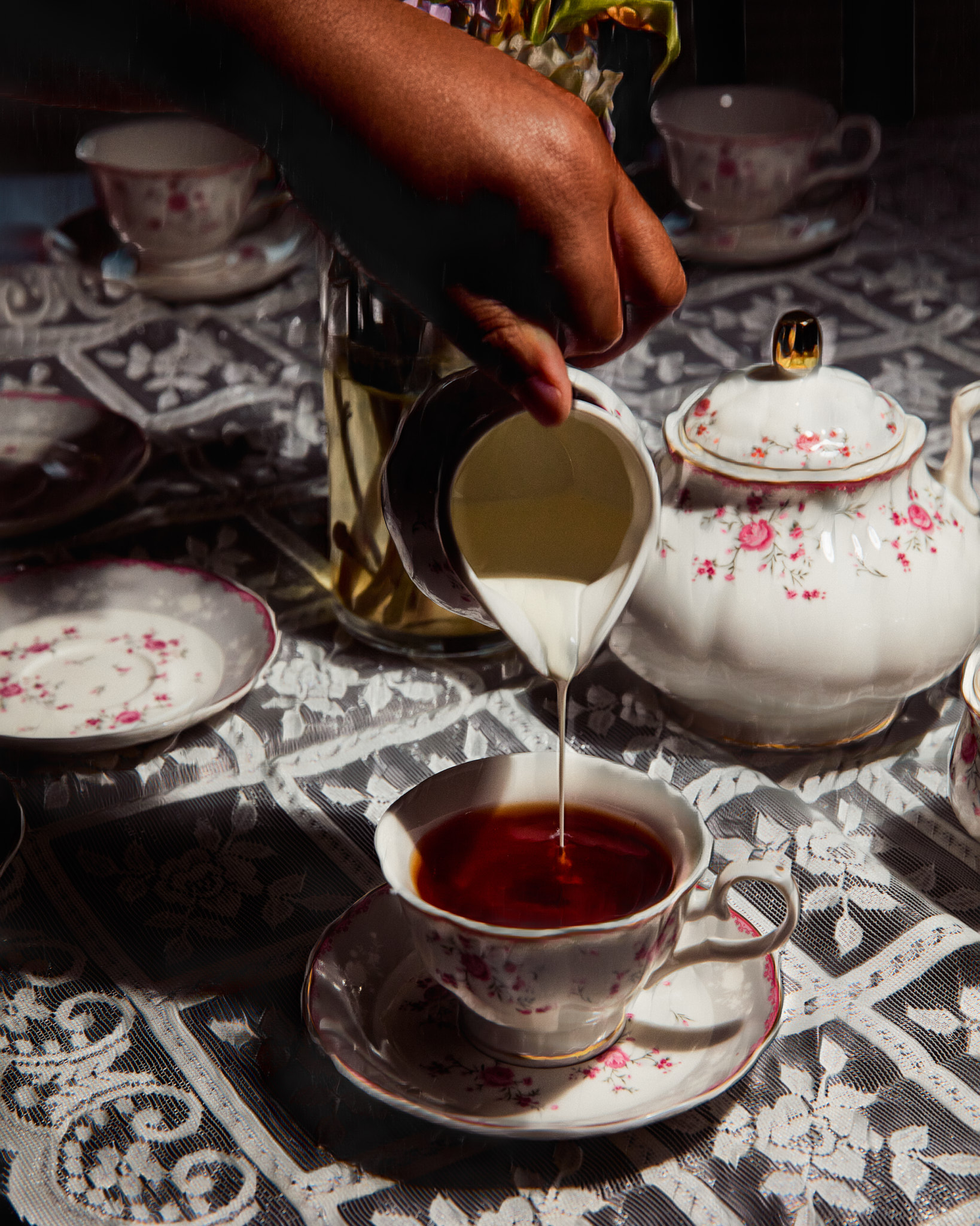 A hand pouring milk into a cup of tea from a creamer on a table set with floral teacups and a teapot.
