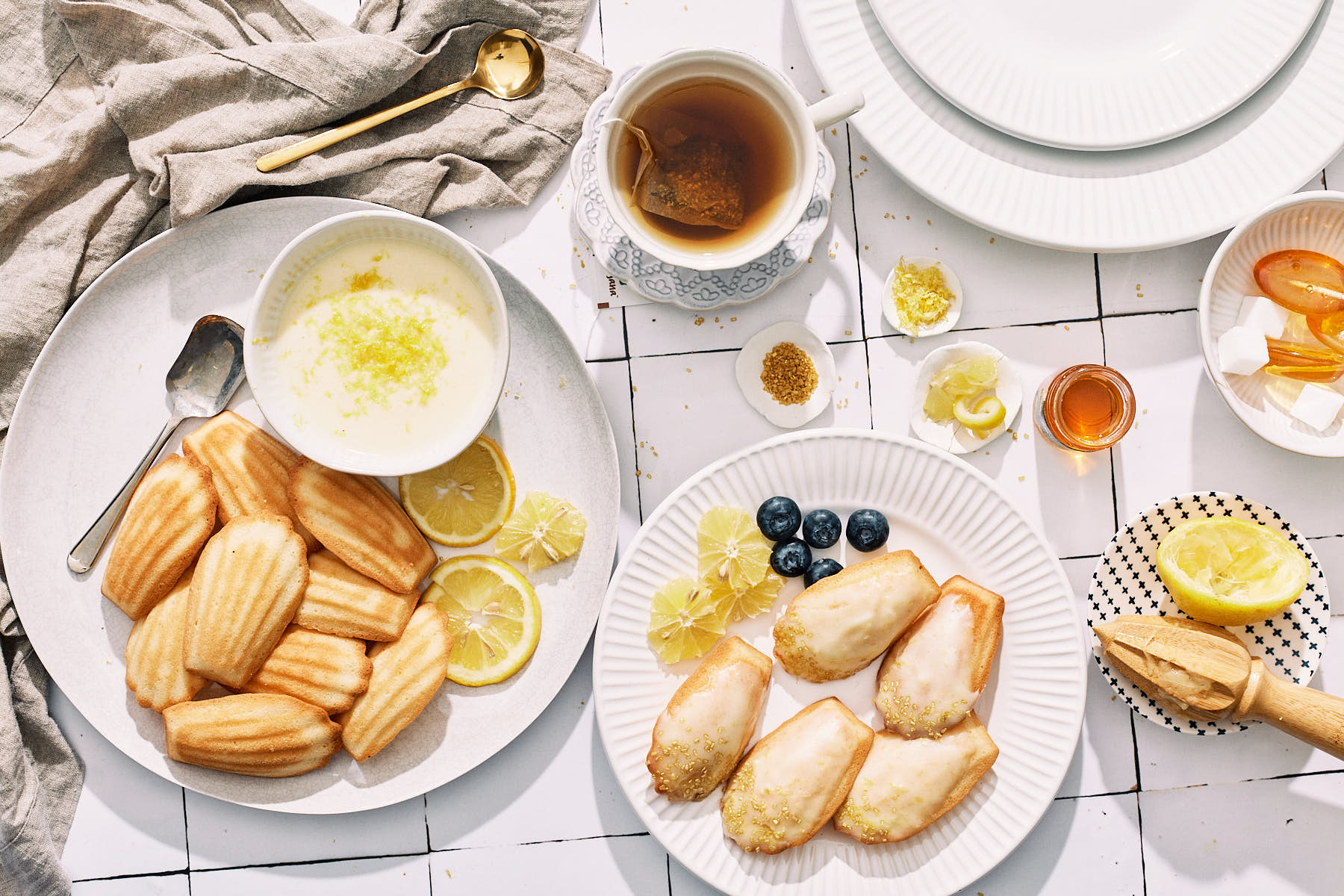 A breakfast table with lemon madeleines, blueberries, tea, lemon curd, and honey on white plates and a white tile surface.