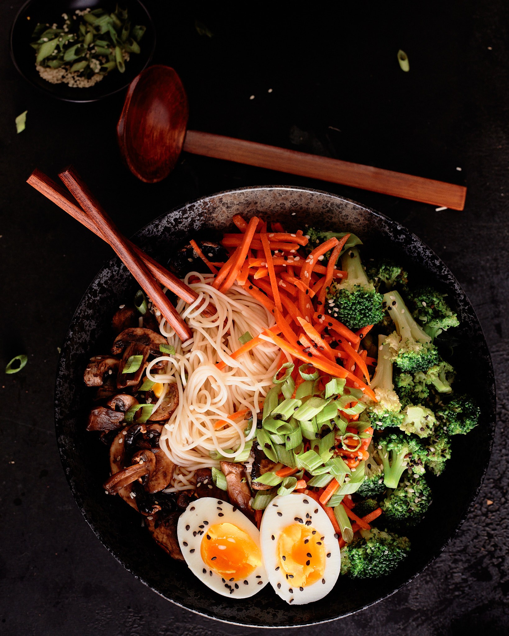 Bowl of ramen with soft-boiled eggs, noodles, broccoli, carrots, mushrooms, and green onions, garnished with sesame seeds, and chopsticks on the side.