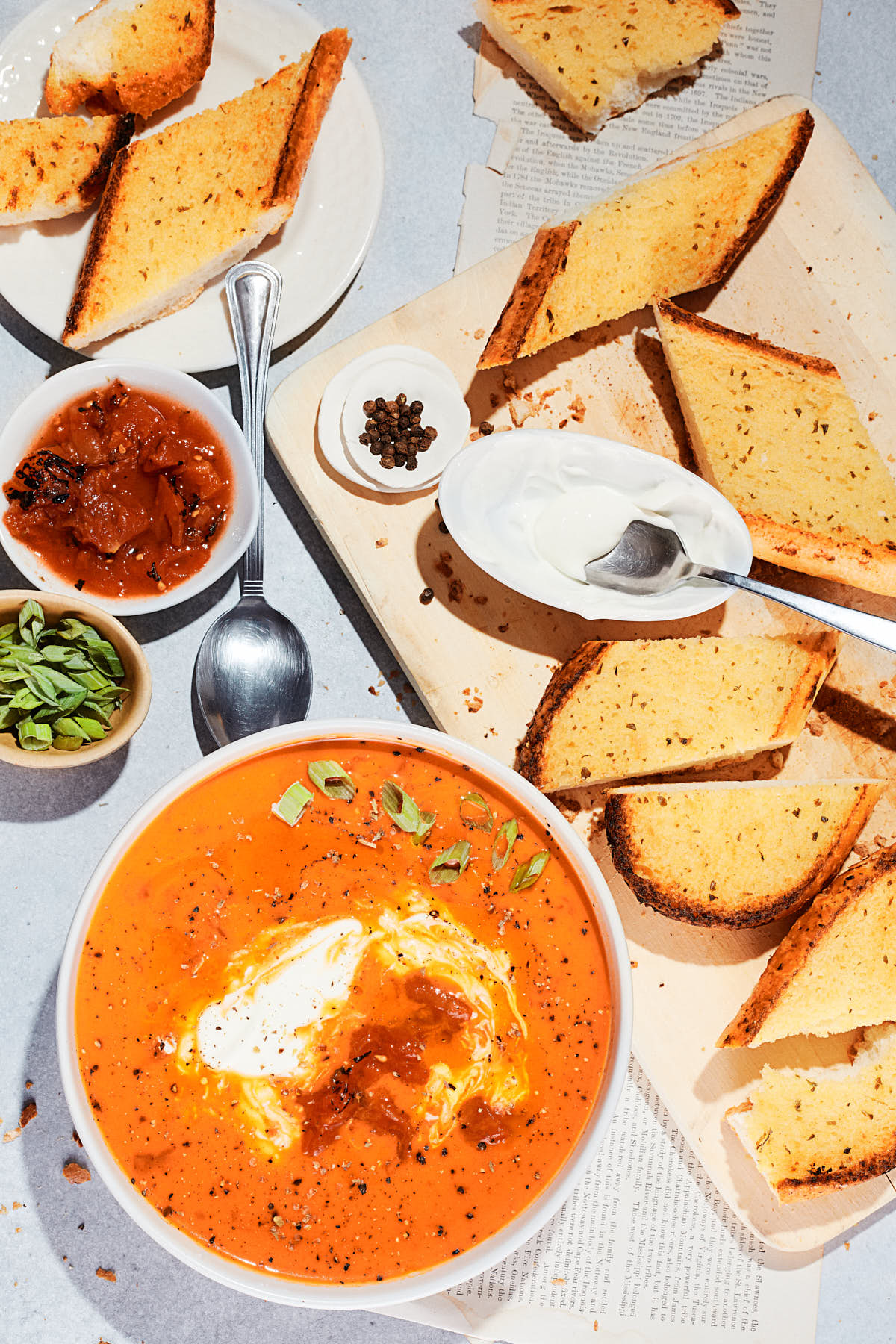 Bowl of tomato soup with sour cream, pepper, and herbs, served with toasted bread slices, chutney, and herbs on a light-colored table.