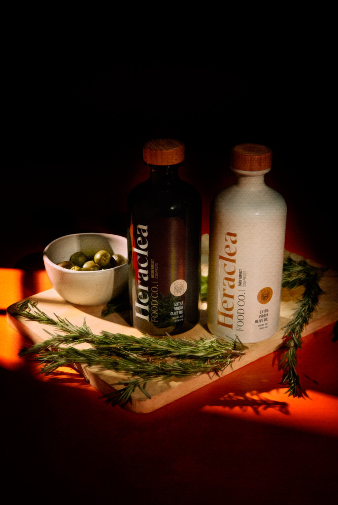 Two Heraclea Food Co. extra virgin olive oil bottles, one black and one white, placed on a wooden board with a bowl of olives and sprigs of rosemary, under warm, dramatic lighting.