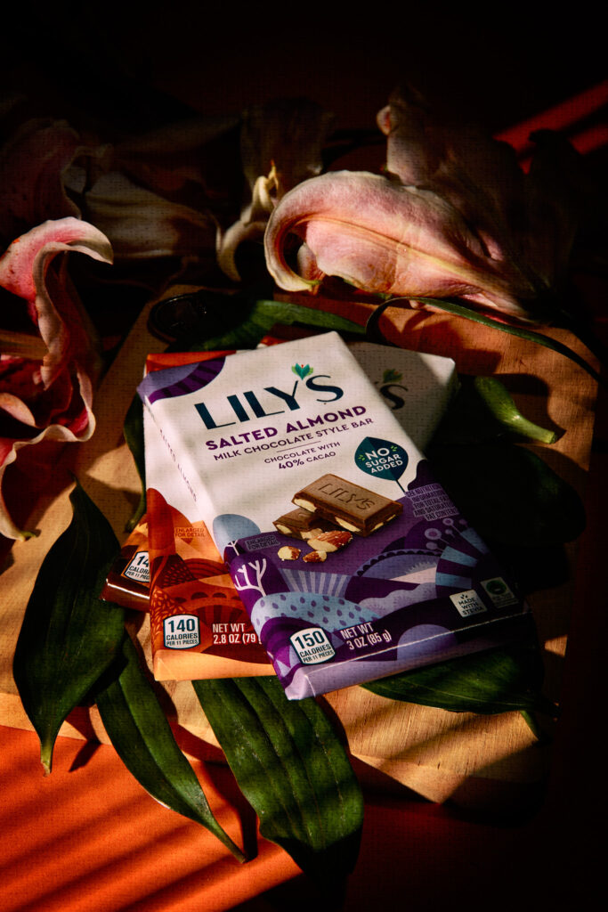 A close-up of two of Lily's salted almond milk chocolate bars is placed on a wooden surface surrounded by pink lilies and green leaves, with dramatic shadows and warm lighting.