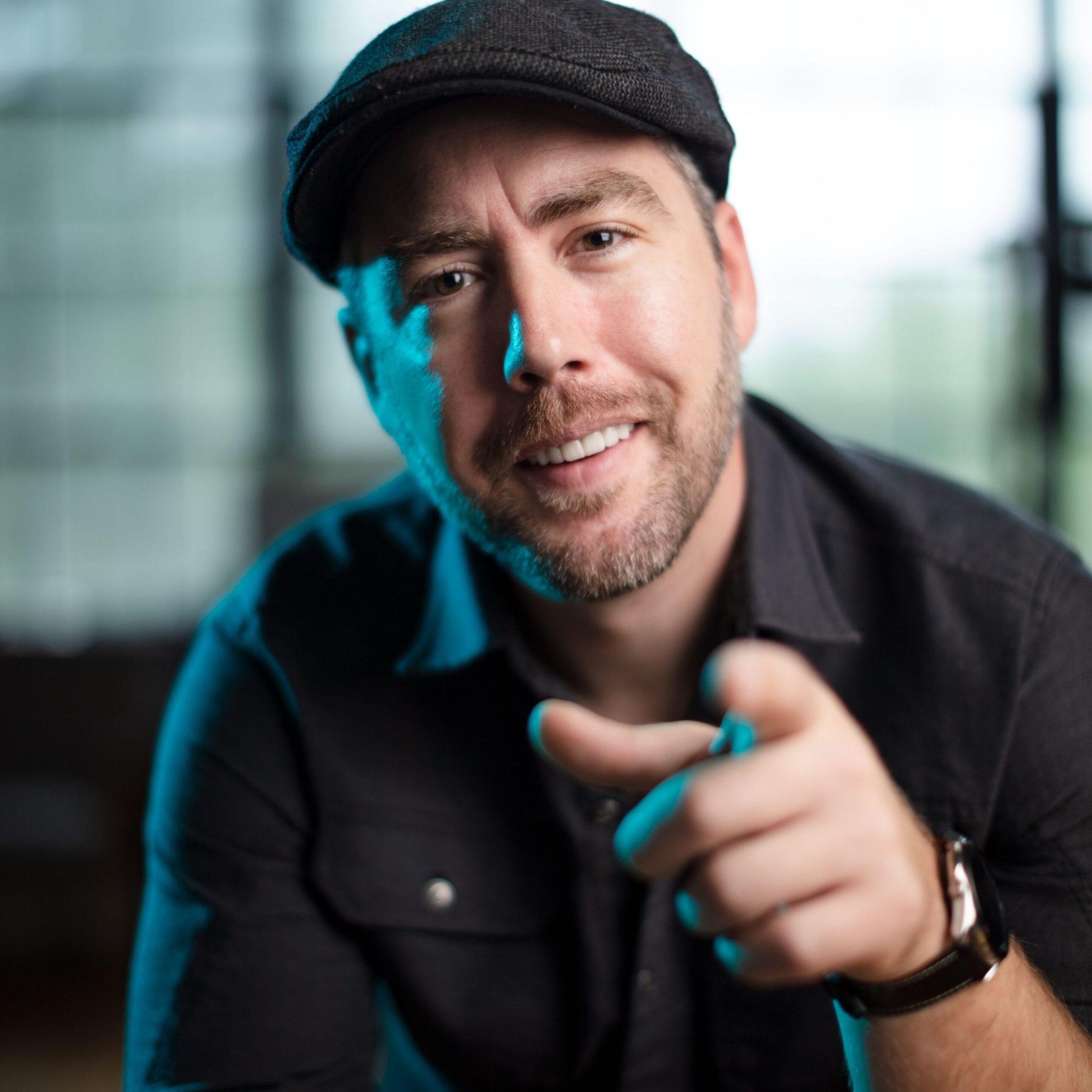 A smiling man wearing a black shirt and a flat cap, pointing towards the camera. The background is softly blurred, creating a focus on his friendly expression.