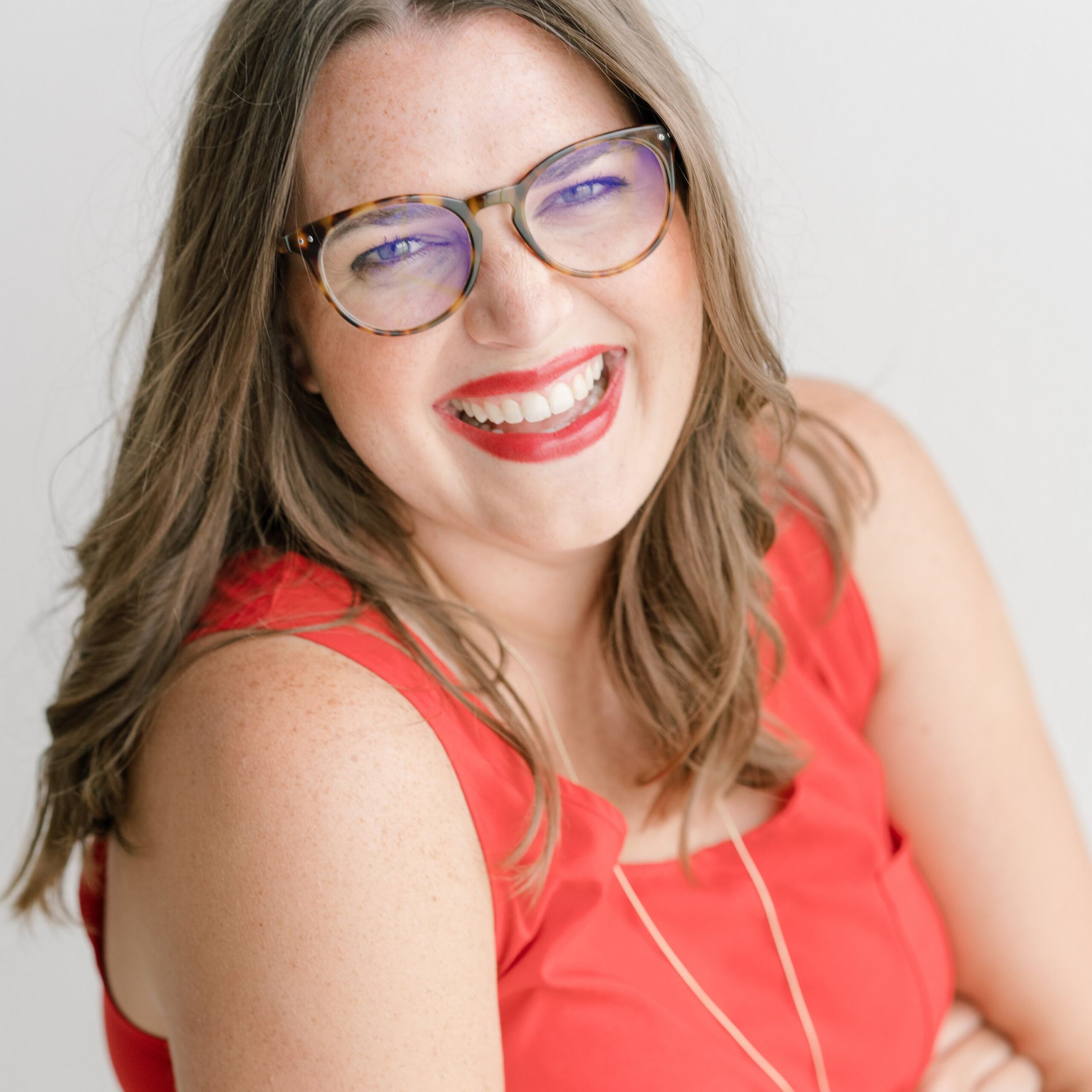 A smiling woman with long brown hair, wearing tortoiseshell glasses and a bright red sleeveless top, poses against a light background.