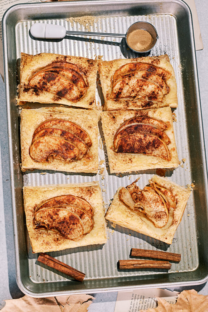 Six apple puff tarts topped with cinnamon arranged on a baking sheet with cinnamon sticks and sugar, creating a rustic dessert scene.