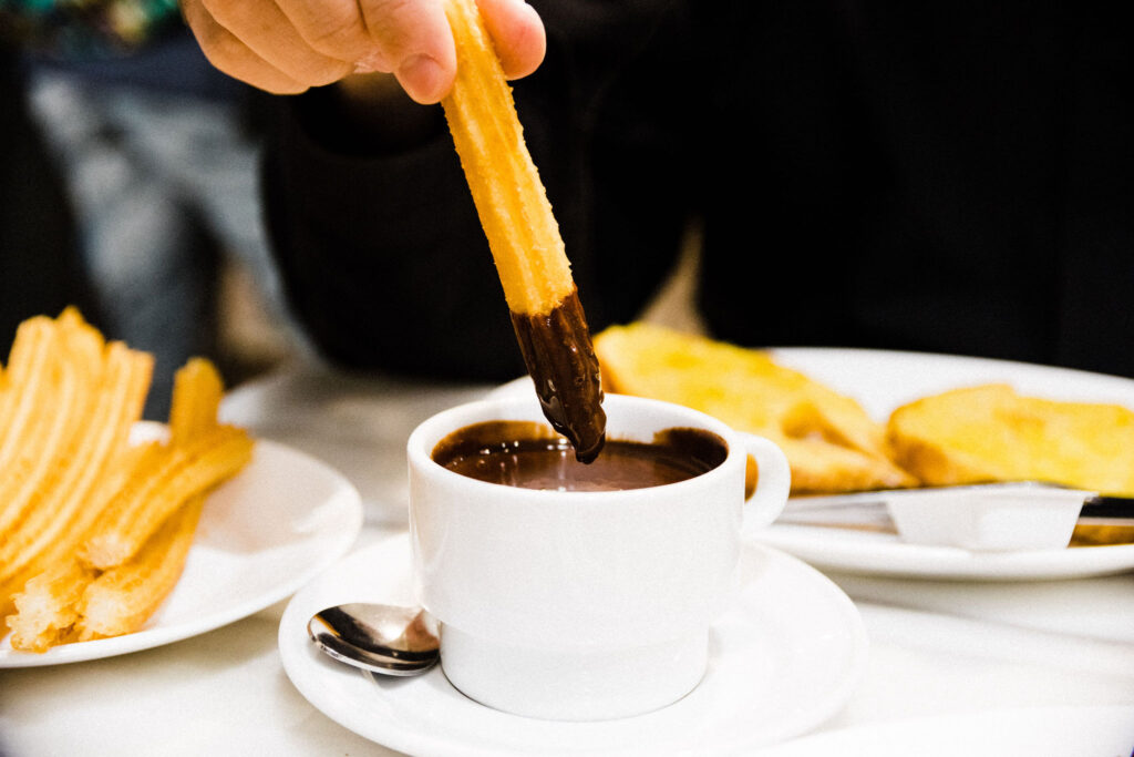 A person dips a churro into a cup of thick hot chocolate on a white saucer, with churros and toasted bread on plates in the background.