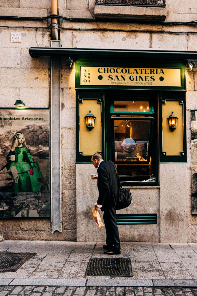 A man in a suit stands outside Chocolatería San Ginés, holding pastries and a cup of coffee, with the shop's green and yellow signage in the background.