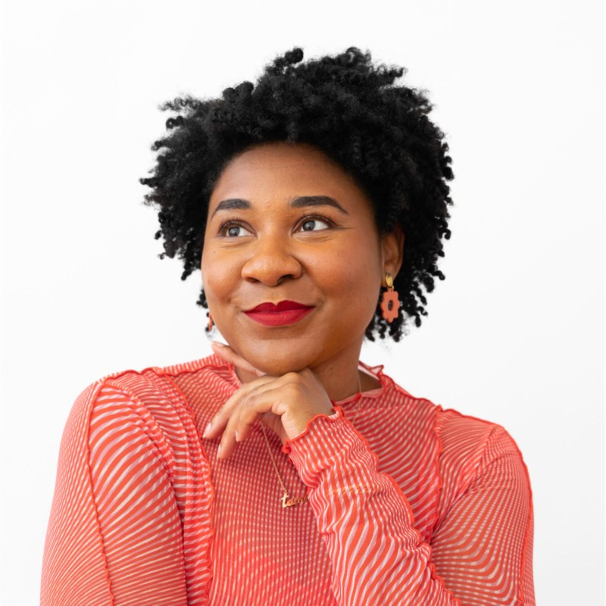 A confident woman with natural curly hair poses against a white background. She wears a red, striped blouse, red lipstick, and orange floral earrings. She rests her chin lightly on her hand, smiling thoughtfully.