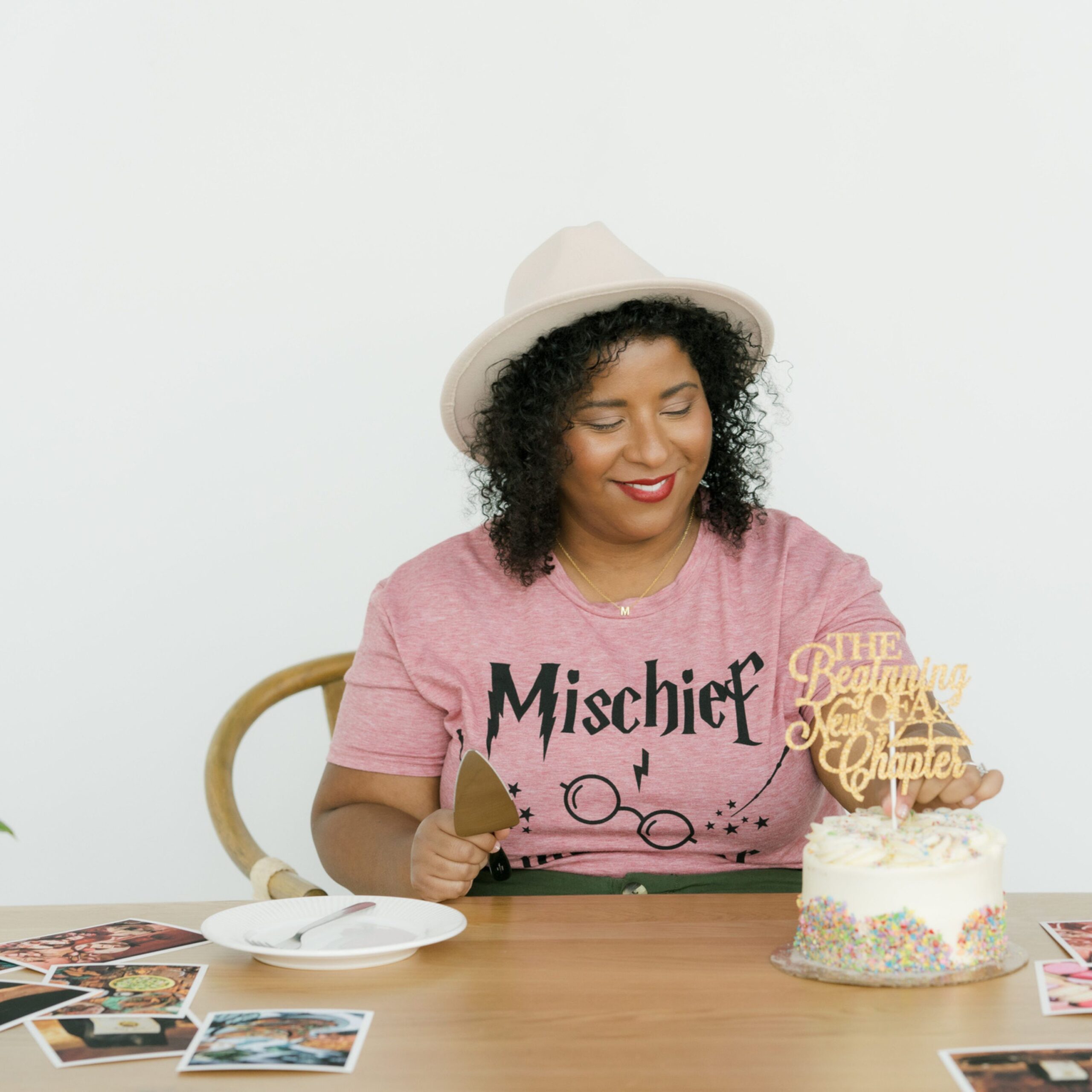 A smiling woman with curly hair wearing a pink hat and a pink “Mischief” T-shirt sits at a wooden table. She holds a cake server and is about to slice a white cake topped with rainbow sprinkles and a gold cake topper reading “The Beginning of a New Chapter.” Several photographs are spread out on the table around her.