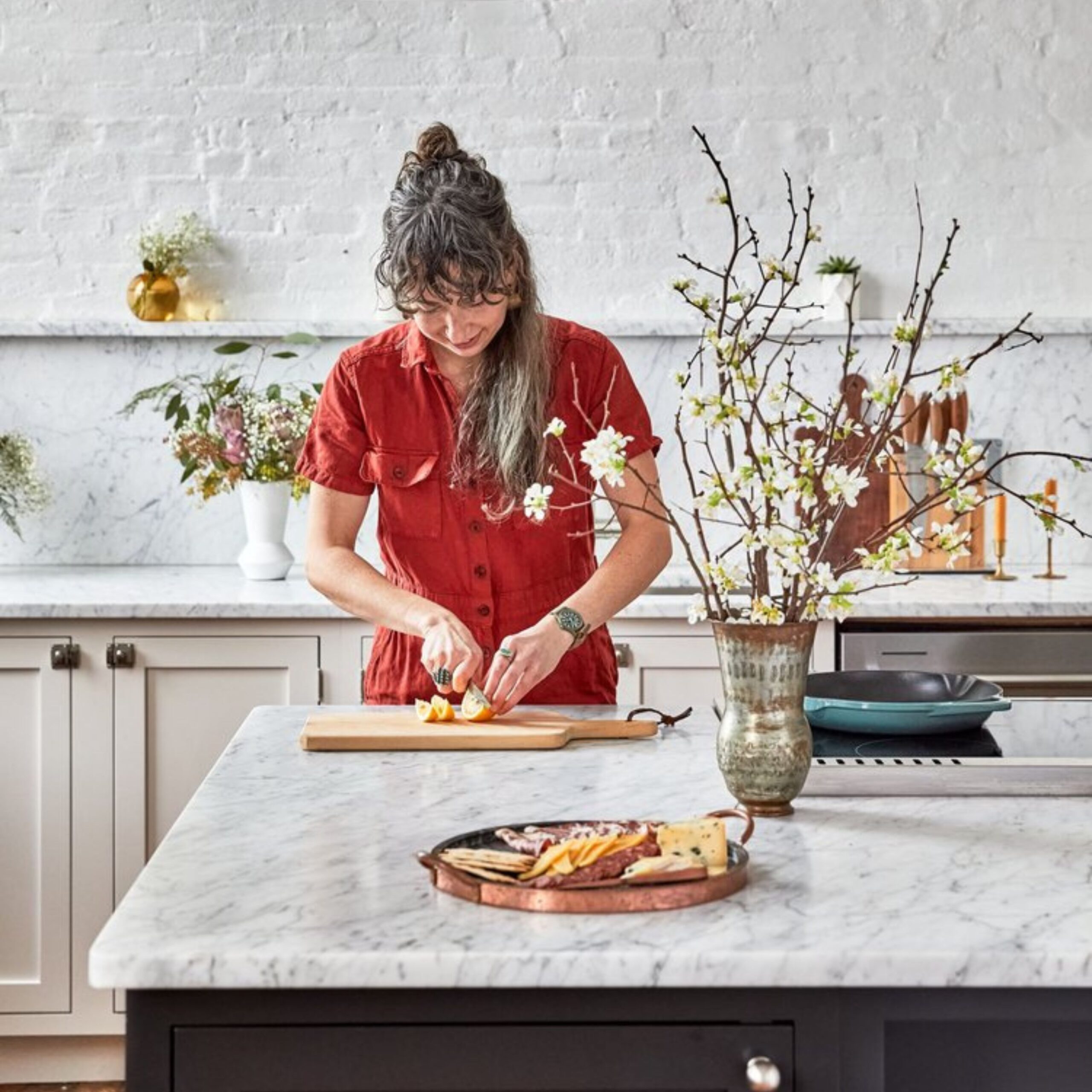 A woman in a red dress slices citrus on a wooden cutting board in a bright kitchen with a marble countertop, surrounded by spring flowers and a cheese platter in the foreground.