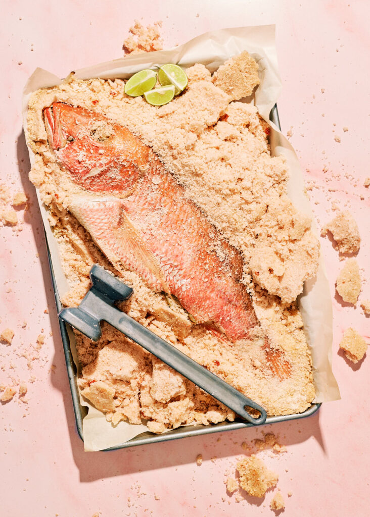 Salt-crusted red snapper partially cracked open on a parchment-lined baking sheet, with a metal mallet resting on the coarse salt and lime wedges as garnish. The pink-hued background complements the golden crust and the fish’s vibrant color.