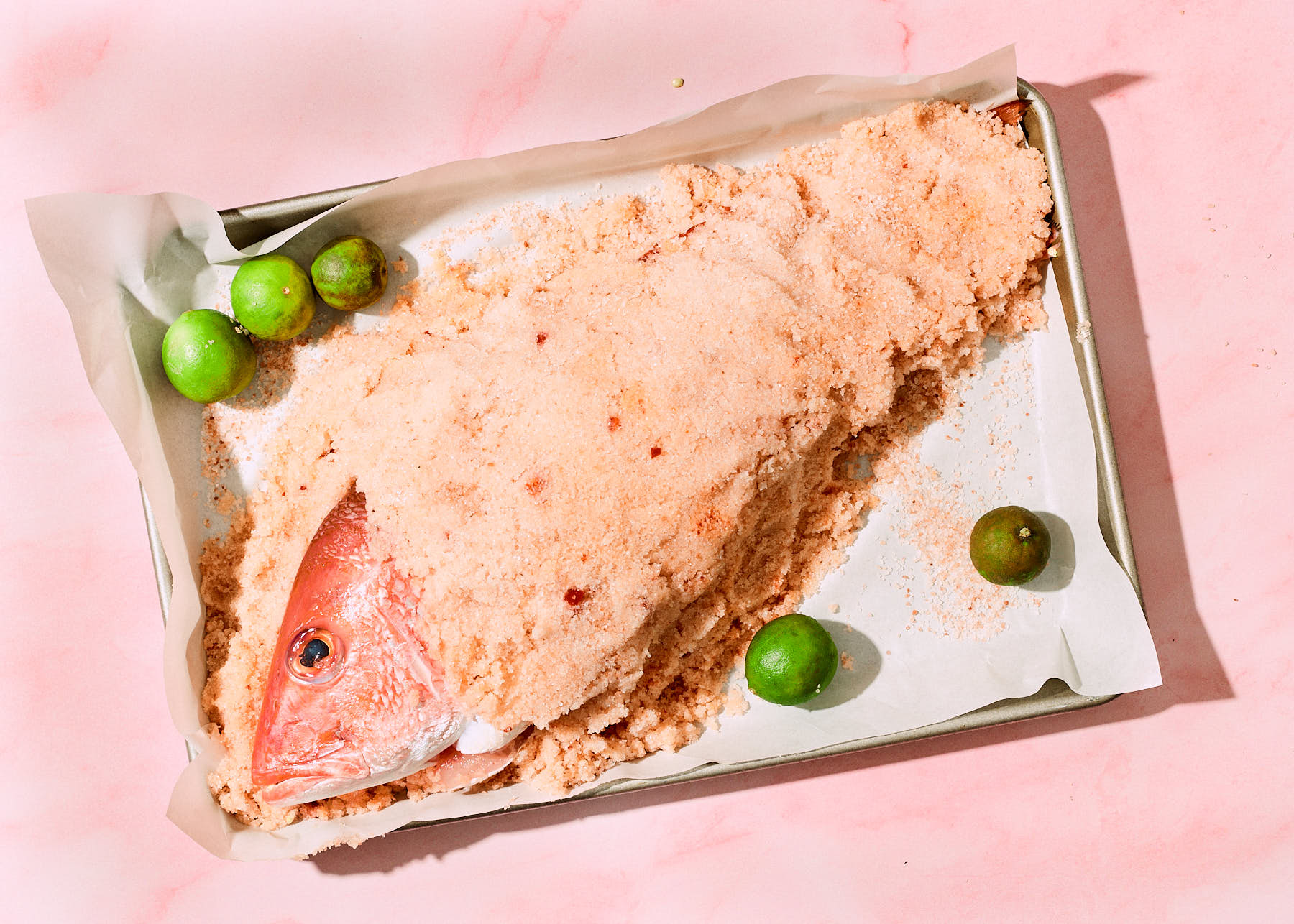 Whole red snapper encased in a thick salt crust on a parchment-lined baking sheet, surrounded by fresh key limes. The pink background enhances the warm tones of the fish and golden salt mixture.