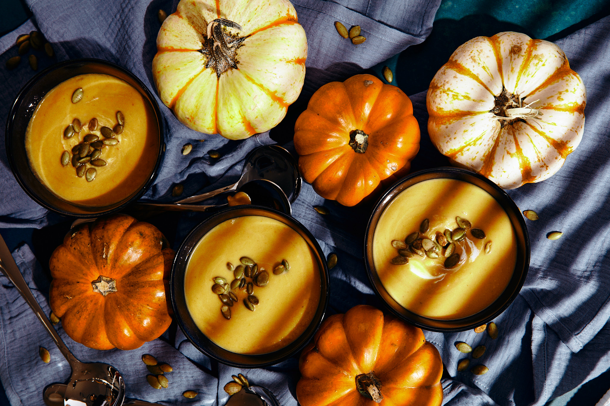 Three bowls of pumpkin soup topped with pepitas surrounded by mini pumpkins on a textured blue cloth.