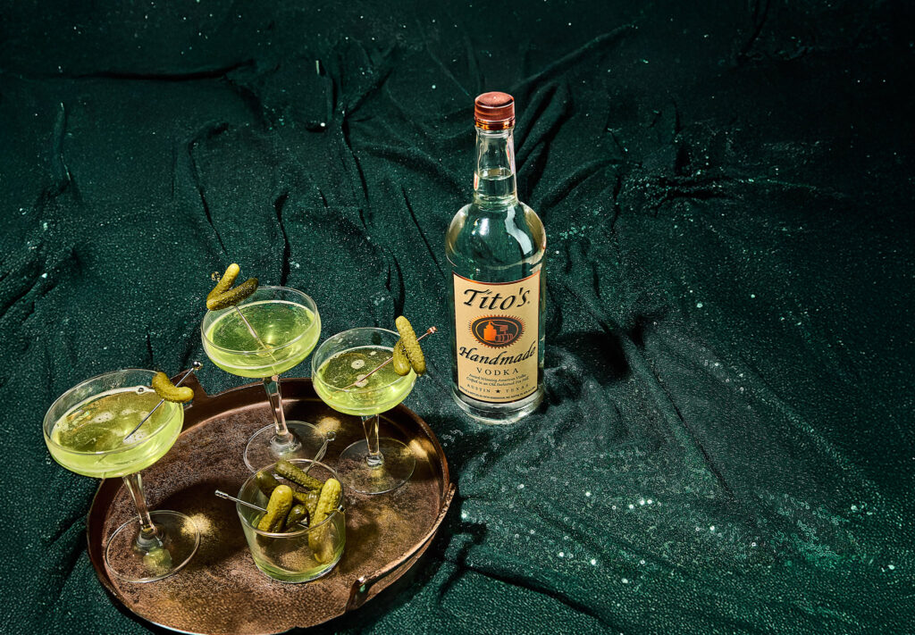 Cocktail photography featuring Tito’s Handmade Vodka pickle martinis on a textured green background, styled with pickles and vintage coupe glasses—shot by Austin food and beverage photographer.