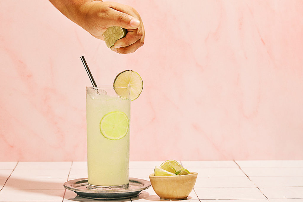 hand squeezing lime juice into agua frescadrink with lime in front of pink backdrop