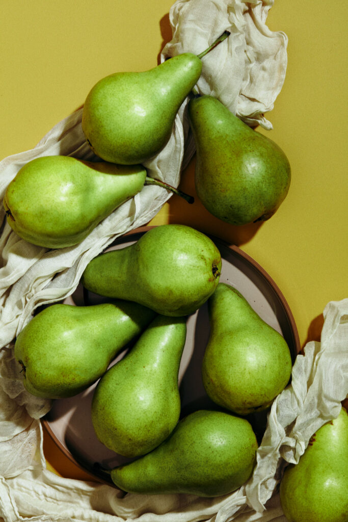 Lush green pears artfully arranged on and around a plate, interspersed with crinkled white cloth, against a mustard yellow backdrop.