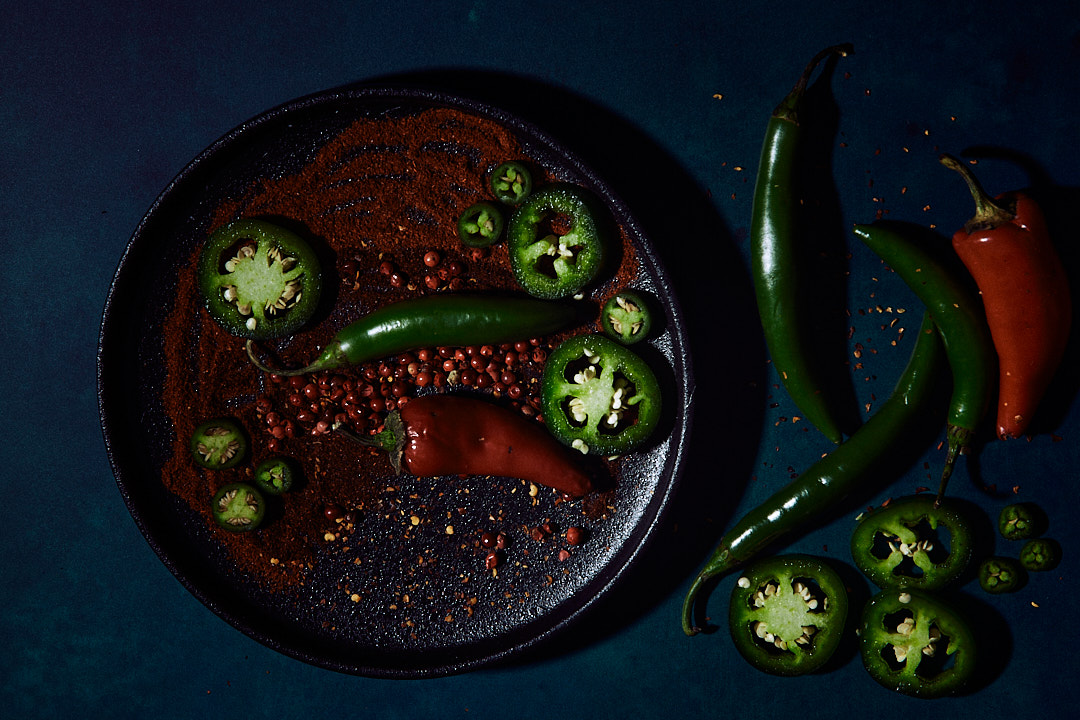 A dark plate featuring sliced green and red chili peppers, with scattered red peppercorns and chili powder against a deep blue background.